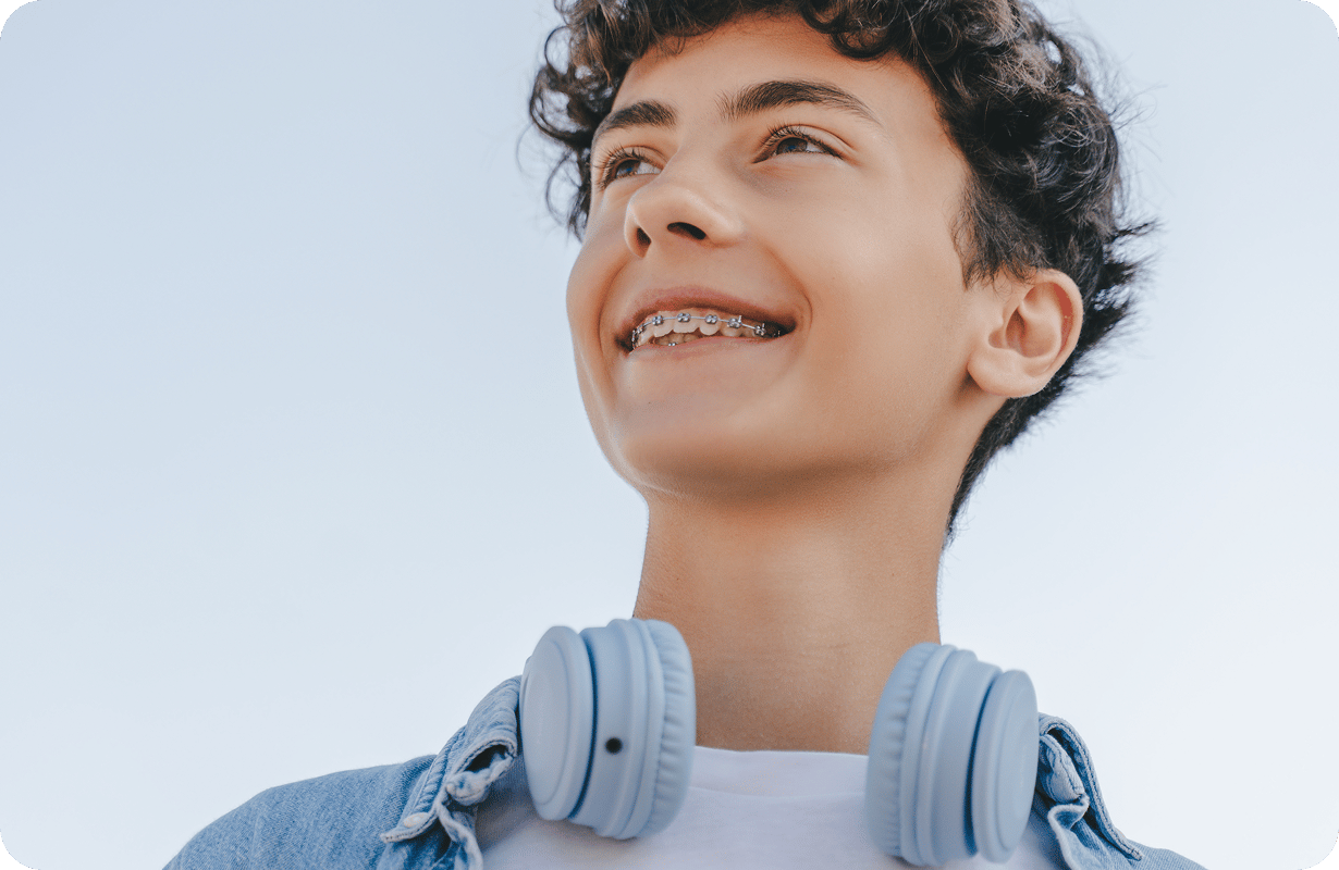 A low-angle portrait of a happy teenage boy with curly hair smiling and showing his metal dental braces.