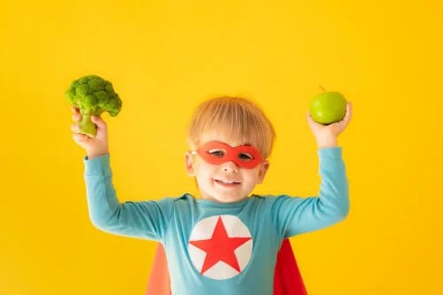 Superhero kid with cape and mask holding broccoli and apple Superhero kid with cape and mask holding broccoli and apple