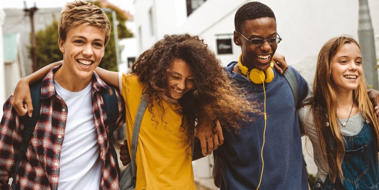 A group of four happy teenagers walking together outdoors, smiling and laughing, with arms around each other's shoulders. A group of four happy teenagers walking together outdoors, smiling and laughing, with arms around each other's shoulders.