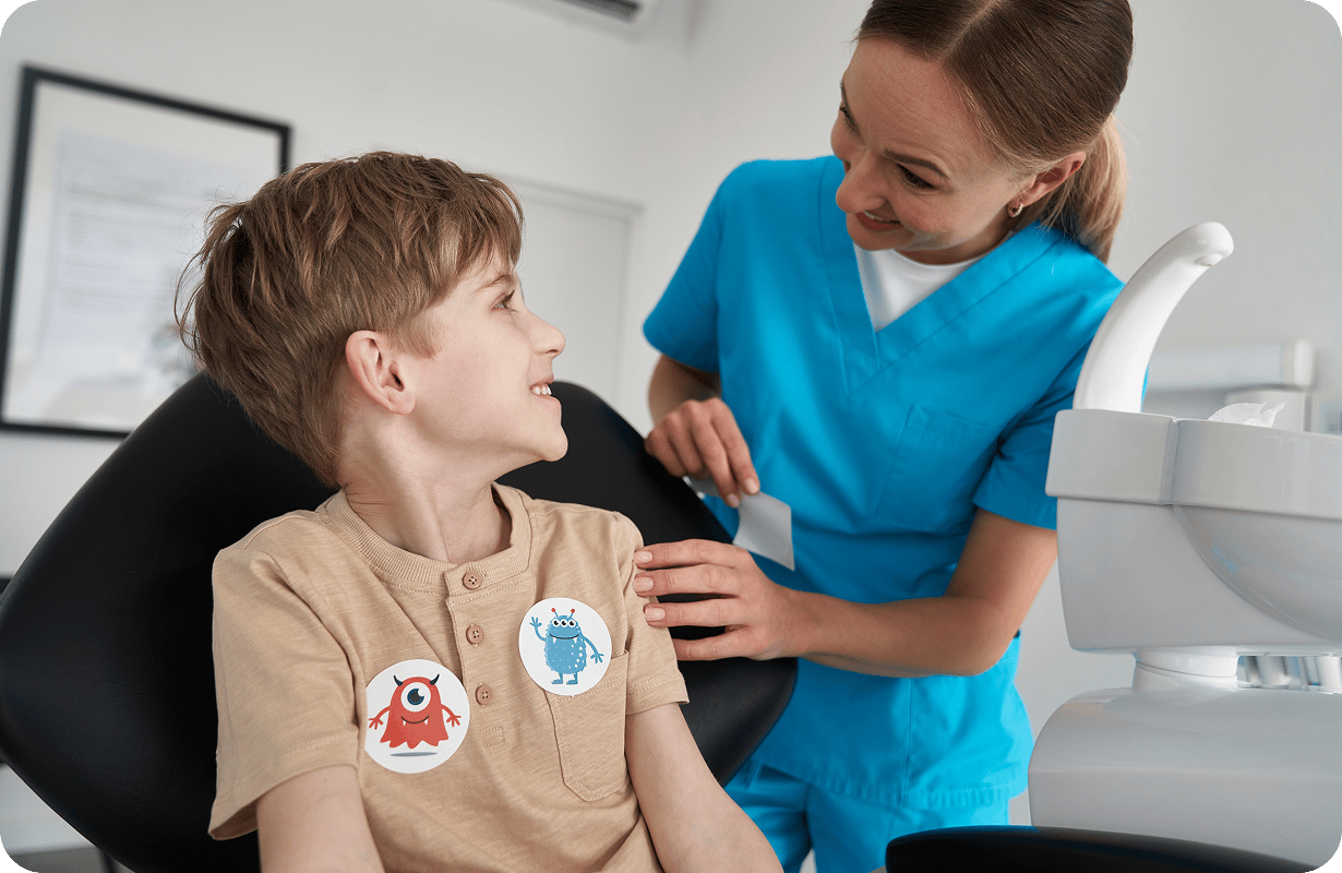 A smiling pediatric dentist in blue scrubs interacting with a young boy sitting in a dental chair.