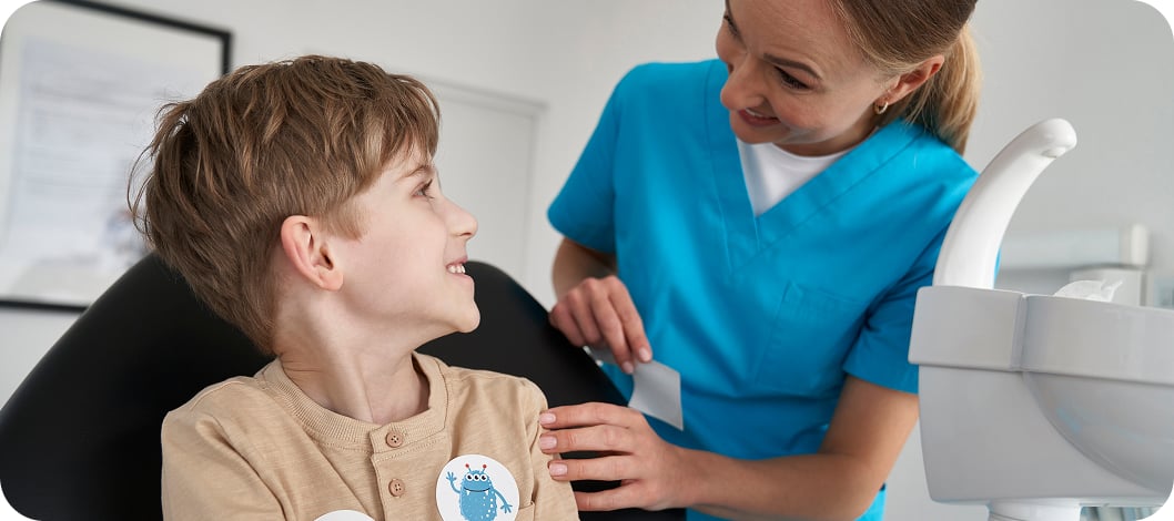 A smiling pediatric dentist in blue scrubs interacting with a young boy sitting in a dental chair.